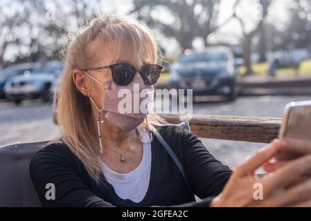 Mature adult woman smiling with glasses wearing face mask looking at smartphone. Stock Photo