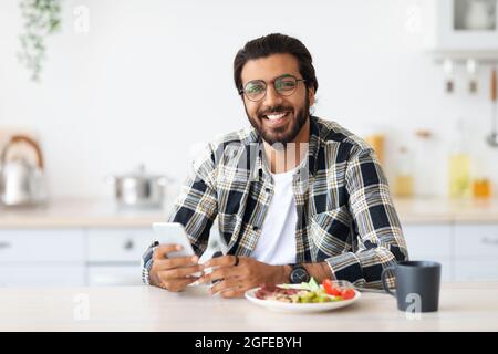 Cheerful middle-eastern man enjoying breakfast, holding smartphone Stock Photo