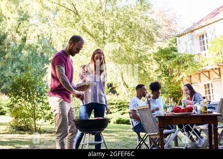Young man preparing food in barbecue by his friends sitting at table Stock Photo