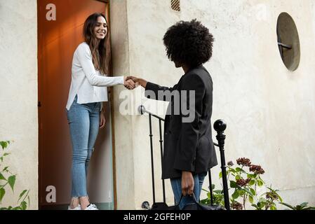 Smiling young female friends shaking hands at doorstep Stock Photo
