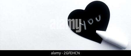 Banner with black heart as a chalkboard on which the chalk says 'YOU', a chalk lies next, the background of a white textured sheet of paper. Copy spac Stock Photo