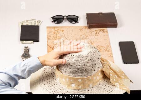 female hand holds a straw hat. on a white wooden table lies a world map, a black wallet with money, a diary, a watch, a smartphone and sunglasses. sel Stock Photo