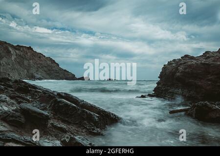 rocky beach landscape during storm with cloudy sky. Dramatic long exposure of sea waves breaking on rocks. Stock Photo
