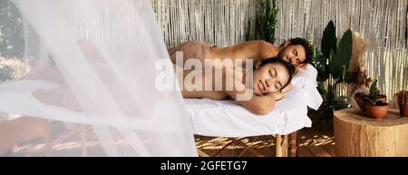Multi-ethnic couple having relax during back massage lying with eyes closed on massage tables at romantic spa resort, summer outdoor. Side view Stock Photo
