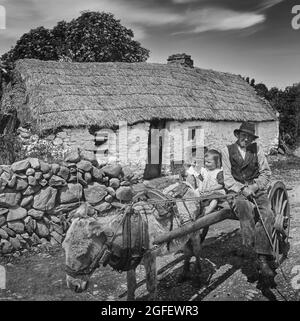 Early 20th century view of a farmer taking children on a donkey cart, whilst his wife watches from the stable door of a traditional thatched on the West Coast of Ireland Stock Photo