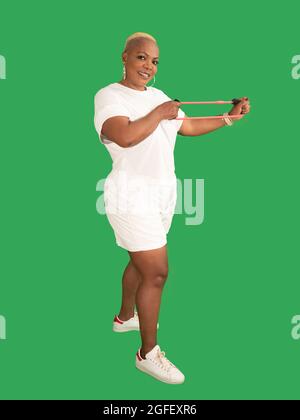 Vertical portrait of a smiling beautiful Afro American woman wearing sportswear and working out with a resistance band isolated against a green backgr Stock Photo
