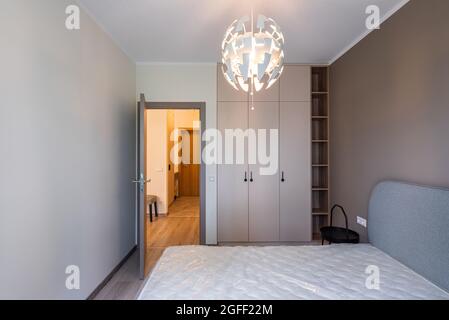 Modern interior of renovated bedroom in apartment. Beige wall. Bed with new white mattress. Black night table. Wooden wardrobe. Opened door to hall. Stock Photo