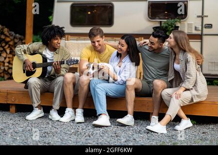 Young diverse friends with pet dog and guitar playing music, having fun near RV on autumn weekend, camping together Stock Photo