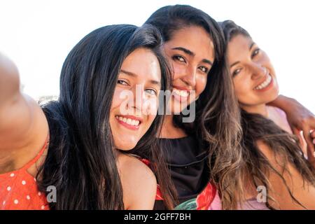 Three latin friends taking a picture of themselves outdoors in a sunny day. Happy hispanic girl friends smiling at camera taking a photo for her socia Stock Photo