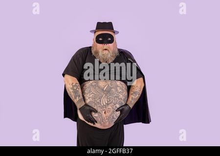 Doubting man with overweight in carnival suit holds huge belly on purple background Stock Photo