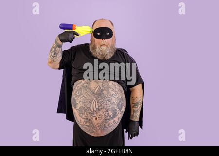 Funny obese man in hero costume holds toy blaster to temple on purple background Stock Photo
