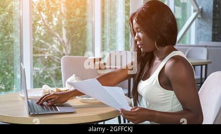 African-American lady looks at papers working on laptop Stock Photo