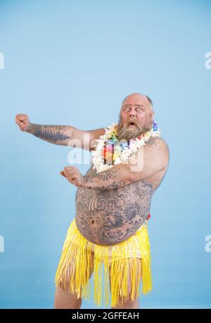 Emotional man with overweight in decorative grass skirt adances on light blue background Stock Photo