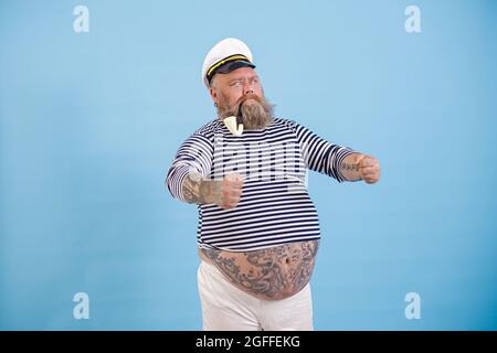 Obese captain with vintage smoking pipe holds invisible helm on light blue background Stock Photo