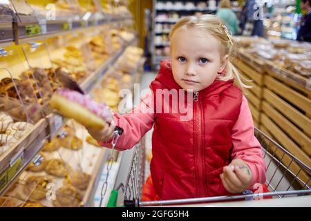 3 years old girl in shopping cart holds donute in marketplace. Stock Photo