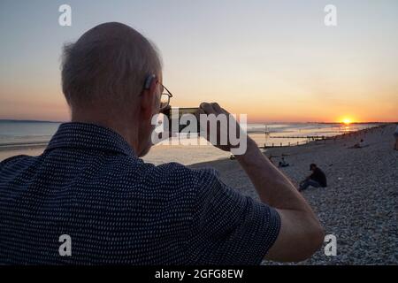 UK weather, 25 August 2021: After a sunny day, clear skies make for beautiful colours in the sky, reflected on the wet sand, as the sun sets over the Solent. A man in his late 70s, wearing a hearing aid, tkes a photo of the sunset on his smart phone. Anna Watson/Alamy Live News