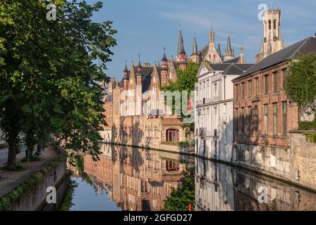 Brugge, Flanders, Belgium - August 4, 2021: Belfry towers over the sunlighted facades of quiet Groenerei canal water mirroring the houses. Some green Stock Photo