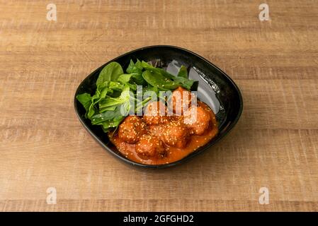 Large serving of minced meat meatballs with tomato sauce and spinach leaves on a shiny black plate. Stock Photo