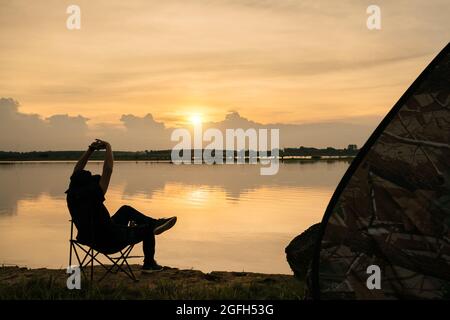 Asian young man traveler relax, enjoying sunset scenery view of the river landscape, sitting in touristic chair. Travel camping and adventure lifestyl Stock Photo