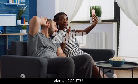 Joyful interracial couple taking pictures on smartphone while sitting in living room. Mixed race husband and wife feeling cheerful taking fun selfies and enjoying happy relationship Stock Photo