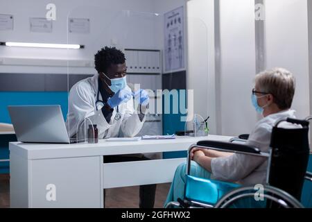 Disabled senior patient discussing illness with black medic in medical office. African american doctor consulting elder woman sitting in wheelchair with healthcare issues and disease Stock Photo