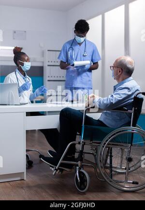 Professional african american team discussing healthcare diagnosis with disabled old patient. Black doctor and nurse talking to sick elder man with handicap sitting in wheelchair Stock Photo