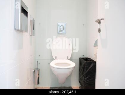 White water closet, restaurant bathroom, small toilet room with a wall mounted, wall hung ceramic toilet bowl, toilet brush, waste basket, toilet pape Stock Photo