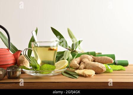Cup of green tea with ginger mint and lemon on wooden table with leaves, metal teapot and bamboo canes on wooden table and white isolated background. Stock Photo