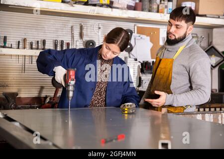 Professional worker helps a female apprentice drill holes in metal plate Stock Photo