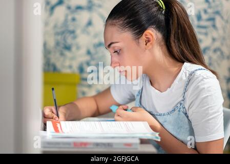 Side view of focused smart teen girl writing in copybook while preparing homework task at table at home Stock Photo