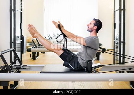 Fit muscular man doing a teaser pilates exercise on a reformer in a gym to strengthen his core and abdominal muscles in a side view with copyspace Stock Photo