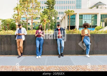 Four young friends wearing masks and social distancing during Covid-19 pandemic standing in a line in the street leaning against a low wall engrossed Stock Photo