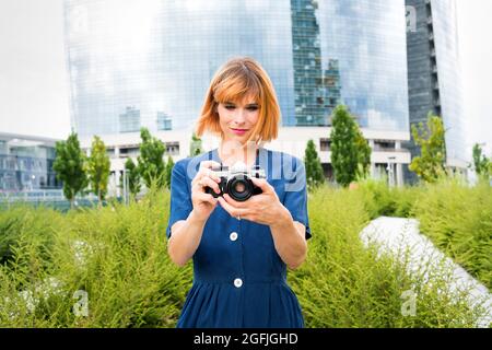 Female tourist using her camera in the city pointing at the camera with a quiet smile as she composes her shot against a backdrop of a modern glass sk Stock Photo