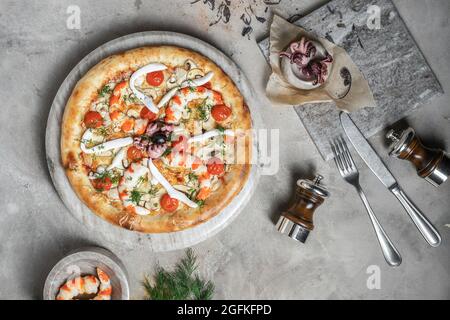 Tasty pizza with  tuna, seafood, shrimps and mini octopus on marble round tray. Garnished on grey stone backdrop with pizza ingredients Stock Photo