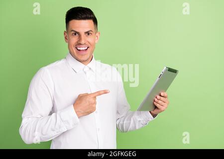 Photo of adorable excited young guy dressed white shirt pointing modern device open mouth smiling isolated green color background Stock Photo