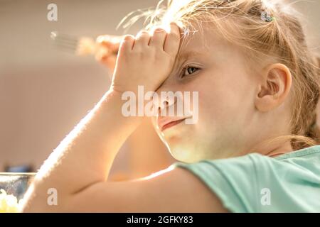 Portrait of a little blonde girl in the kitchen with a fork in her hands. Toddler's baby lunch time. Stock Photo