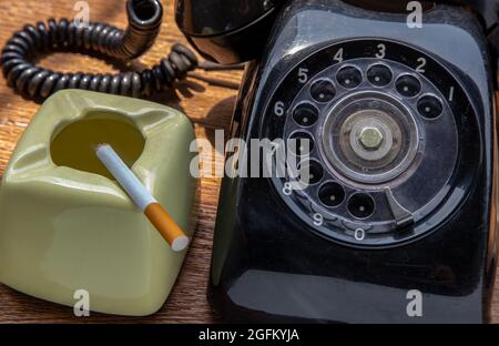 Cigarette that were burning placed on greyish green ashtray and Old black retro rotary telephone on old wooden rustic table. Copy space, Selective foc Stock Photo