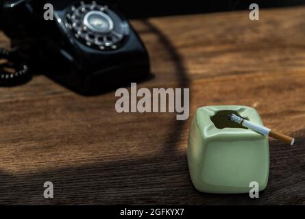 Cigarette that were burning placed on greyish green ashtray in front of Old black retro rotary telephone on old wooden rustic table. Copy space, Selec Stock Photo