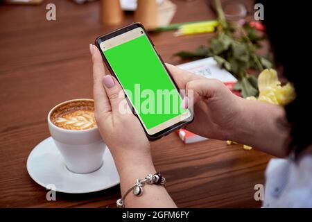 Chroma key mobile phone screen in woman's hand and cup of coffee on the table. Top view. Education, working, freelance concept. High quality photo Stock Photo
