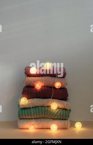 Stack of warm knitted women's sweaters. Garland lights decorations. Winter holidays concept. Stock Photo