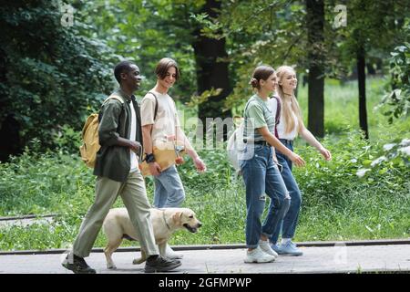 Cheerful teenage girl walking near interracial friends with retriever and skateboard in park Stock Photo