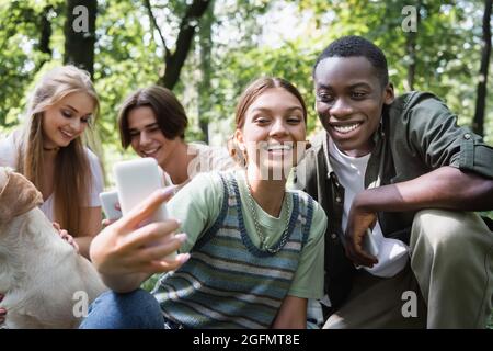 Smiling teenage girl taking selfie near african american boy and friends with retriever in park Stock Photo