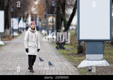 The young woman and empty vertical billboard on the alley in the city park. Empty blank white mockup of billboard. Copyspace. Stock Photo