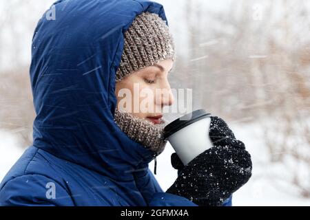 Portrait of a young woman with a cardboard cup of coffee in her hands surrounded by flying snowflakes on the background of a winter forest. Shallow fo Stock Photo