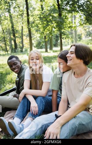 Cheerful teen friends sitting near african american boy with acoustic guitar in park Stock Photo