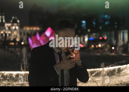 Portrait of young caucasian man in formal black suit lights a cigarette from a lighter. Night picture. Neon signs Stock Photo