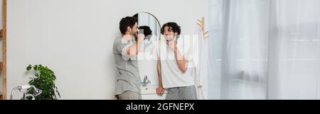 bearded gay couple looking at each other and smiling while brushing teeth in morning, banner Stock Photo