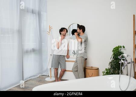 bearded gay couple looking at each other and smiling while brushing teeth in morning Stock Photo