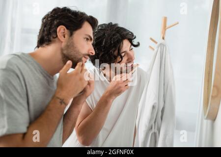 blurred and tattooed gay man touching face while boyfriend brushing teeth and smiling in bathroom Stock Photo