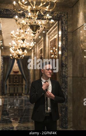 Portrait of young handsome business man in elegant business suit walking on the hall with beautiful expensive chandeliers. straightens his tie